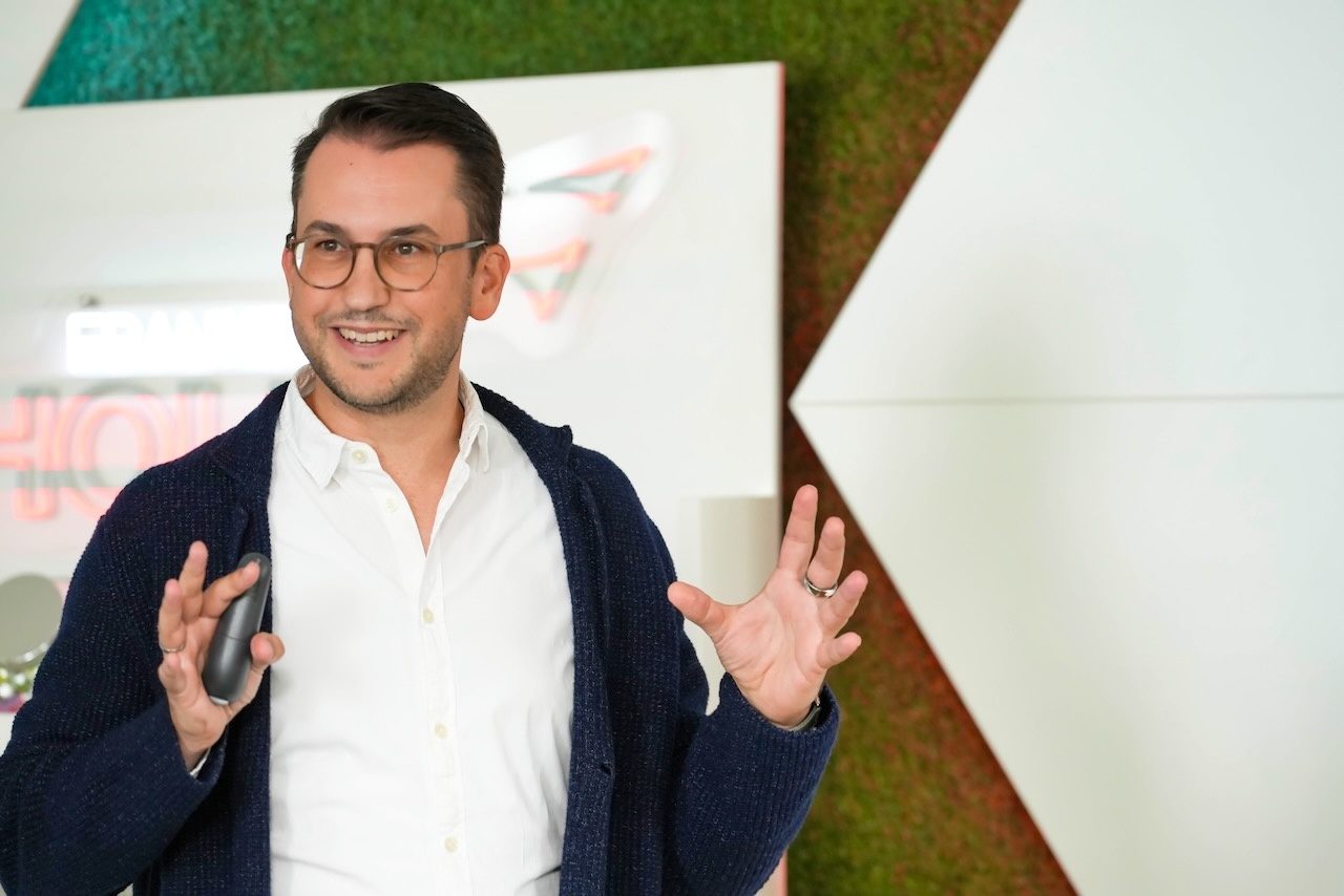 Peter von Aspern steht vor einer Leinwand und erklärt gestikulierend etwas, in der Hand einen Presenter.
